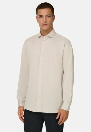 Jeune homme portant une chemise beige à manches longues boutonnée et un pantalon foncé, debout devant un fond clair uni.