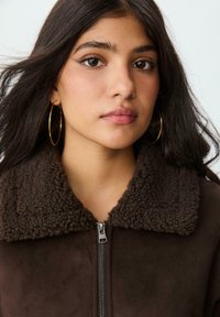 Giacca zip casual marrone con un colletto in shearling soffice, caratterizzata da una consistenza liscia e elementi a contrasto. Orecchini a cerchio dorati accentuano il look.