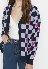 Granatowy i lawendowy kardigan w kratę z różowymi serduszkami, dzianinowy materiał, guziki z przodu i ściągacze przy mankietach. Noszony na białej bluzce.