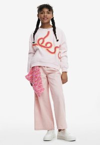 Ragazza con capelli intrecciati che indossa un maglione rosa con un design colorato, pantaloni a gamba larga rosa chiaro, sneaker bianche e tiene una sciarpa di maglia rosa.