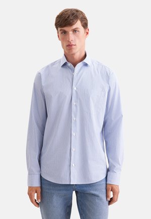 Junger Mann trägt ein hellblaues, langärmeliges Hemd mit dünnen vertikalen Streifen und blaue Jeans vor einfachem Hintergrund.