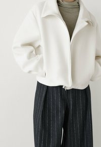 White fleece jacket with a zippered front, high collar, and relaxed fit, paired with dark pinstripe trousers. Soft texture and minimal design.