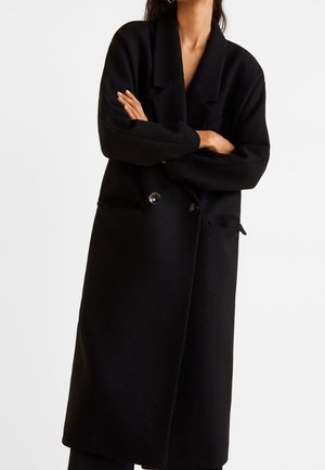 Woman wearing a long black double-breasted coat with wide lapels and front pockets, arms crossed over chest.