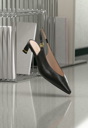 Chaussure en cuir noire à bout pointu avec bride arrière et talon bloc de hauteur moyenne avec accent doré, placée près d'une étagère métallique et de livres sur un sol réfléchissant.