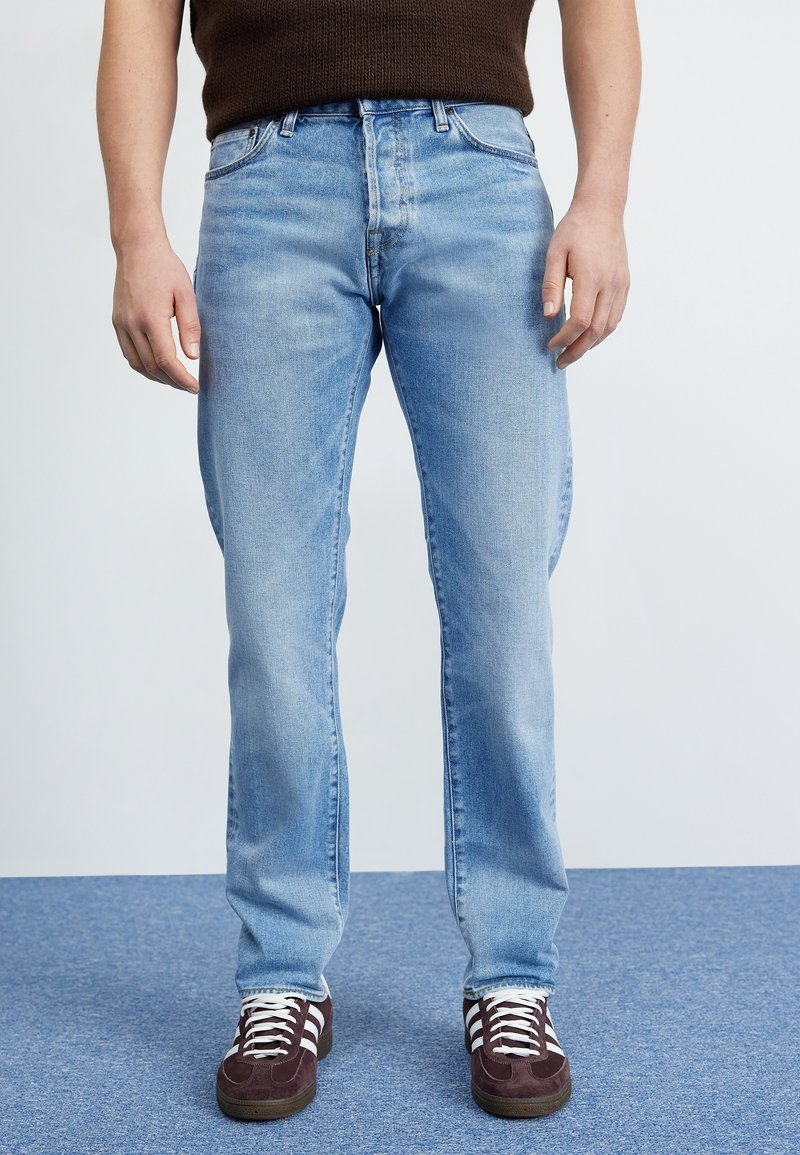 Jeans de mezclilla azul claro con diseño de pierna recta, estilo de cinco bolsillos y un sutil desgaste, combinados con zapatillas de ante marrón oscuro.