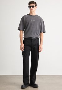 Graues Baumwoll-T-Shirt mit "HUGO"-Logo, kombiniert mit schwarzen Cargo-Hosen mit seitlichen Taschen und schwarzen Plateauschuhen.