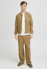 Tan bomberjack met geribde manchetten, twee voorkruipzakken, gecombineerd met brede tan broek en een witte crew neck shirt eronder.