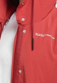 Red lightweight jacket with a hood, featuring a front zipper, snap buttons, and white printed text detail on the chest. Smooth texture.