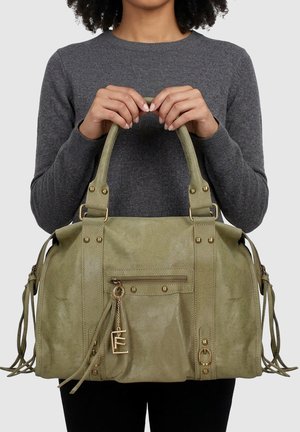 Donna che tiene una grande borsa in pelle verde oliva con tasca frontale con zip e ciondolo dorato, indossa un maglione grigio scuro a manica lunga.