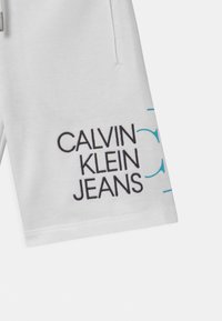 Bílé šortky s černou a tyrkysovou výšivkou a textem „CALVIN KLEIN JEANS“. Hladký bavlněný materiál; včetně bočních kapes.