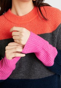 Mikina z pestrých přízí s širokými pruhy v oranžové, šedé, růžové a námořnické modré barvě. Hladká textura s kulatým výstřihem a dlouhými rukávy.