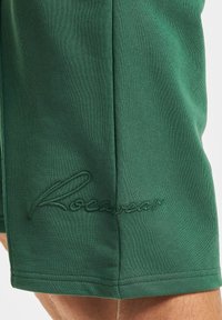 Shorts en coton vert foncé avec une texture lisse, mettant en avant un logo "Rocawear" brodé sur l'ourlet et des détails de couture soignés.