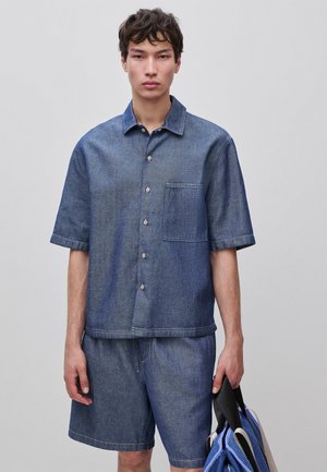 Joven con camisa azul de mezclilla de manga corta abotonada y pantalones cortos a juego, sosteniendo bolsas tote azules y beige en la mano derecha.