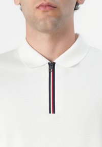 Biała koszulka polo z teksturowanym kołnierzykiem oraz półzamkiem, ozdobiona pionowym niebiesko-czerwonym paskiem. Gładki materiał, minimalistyczny design.