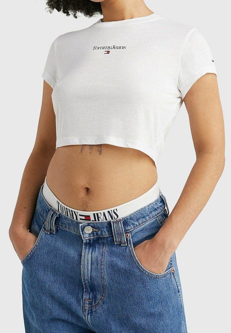 Bílý bavlněný krátký rukáv tričko s logem "Tommy Jeans" v černé a červené barvě, v kombinaci s džínami s vysokým pasem v modré barvě, které mají bílý elastický pas s logem.