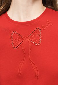 Κοντινό πλάνο του λαιμού κόκκινης μπλούζας με κεντημένο φιόγκο και λεπτομέρειες με ανοίγματα πάνω από το στήθος, με καλλιγραφικό κείμενο "feelin' lucky."