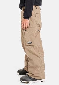Beige snøbrettbukser med løs passform, store sidelommer og en svart logo-lapp. Stoffet ser slitesterkt og vanntett ut.