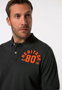 Zwarte poloshirt met een gestructureerde afwerking, voorzien van een oranje geborduurd logo met de tekst "HERITAGE 80" op de linkerkant van de borst. Twee zichtbare knopen.