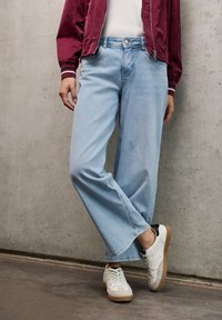 Jean large bleu clair en denim, avec une taille haute et des coutures discrètes. Associé à des baskets blanches et une veste bomber bordeaux.