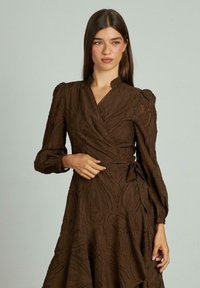 Rochie maro, dintr-un material texturat, cu mâneci bufante și o panglică la talie. Prezintă un tiv ondulat și detalii ornamentale.