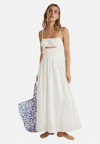 White maxi dress with thin straps, a front cutout, and smocked waist. Accompanied by a patterned blue and white tote bag.