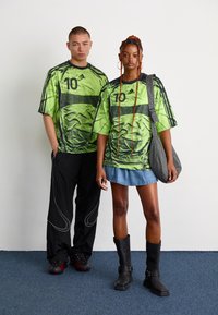 To unge voksne iført matchende grønne Adidas sportsdrakter med nummer 10; en i svarte bukser og joggesko, den andre i en denimskjørt, støvler og skulderveske.