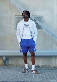 Camisa branca de botões sobre uma t-shirt branca com estampa, calções desportivos azuis com emblema italiano, meias brancas, ténis castanhos, em pé junto a uma parede.