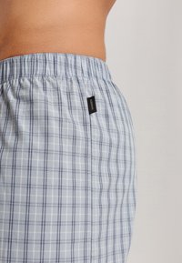 Boxers à carreaux bleu clair avec une taille élastique, dotés de fines lignes navy et d'une petite étiquette noire sur le côté. Texture de tissu douce.