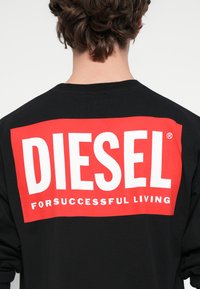 Melna džemperis ar garām piedurknēm, uz muguras liels sarkans taisnstūra logotips ar uzrakstu „DIESEL” baltā krāsā un zem tā „FOR SUCCESSFUL LIVING”.