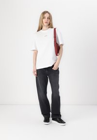 T-shirt blanc en coton avec un petit logo, associé à un jean en denim gris foncé et des baskets noires. La personne tient un sac à bandoulière rouge.