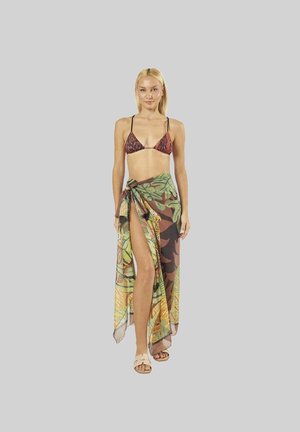 Donna con top bikini fantasia e un sarong colorato con stampa tropicale legato in vita, che cammina in avanti su uno sfondo semplice.