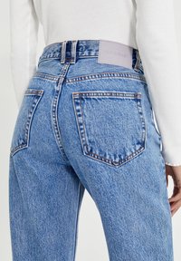 Modré džíny s vysokým pasem a zadními kapsami, se světle fialovou etiketou, nošené s bílým žebrovaným topem s dlouhým rukávem na osobě.