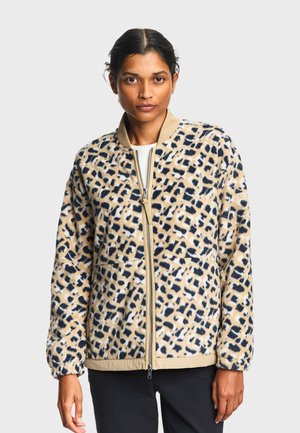 Kvinde iført en beige og sort leopardprintet fleecejakke med lynlås, beige krave og manchetter, stående foran en ensfarvet baggrund.