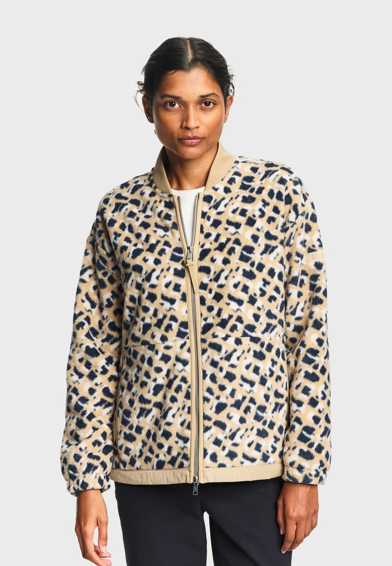 Femme portant une veste polaire zippée à imprimé léopard beige et noir, avec col et poignets beiges, debout devant un fond uni.