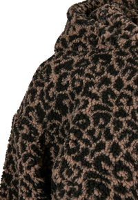 Vêtement en polaire noir et marron à motifs, présentant une surface douce et texturée, avec un col haut et des détails de conception complexes.