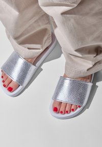 Silver textured slide sandals with a wide strap and a white sole, paired with light beige pants and red-painted toenails.