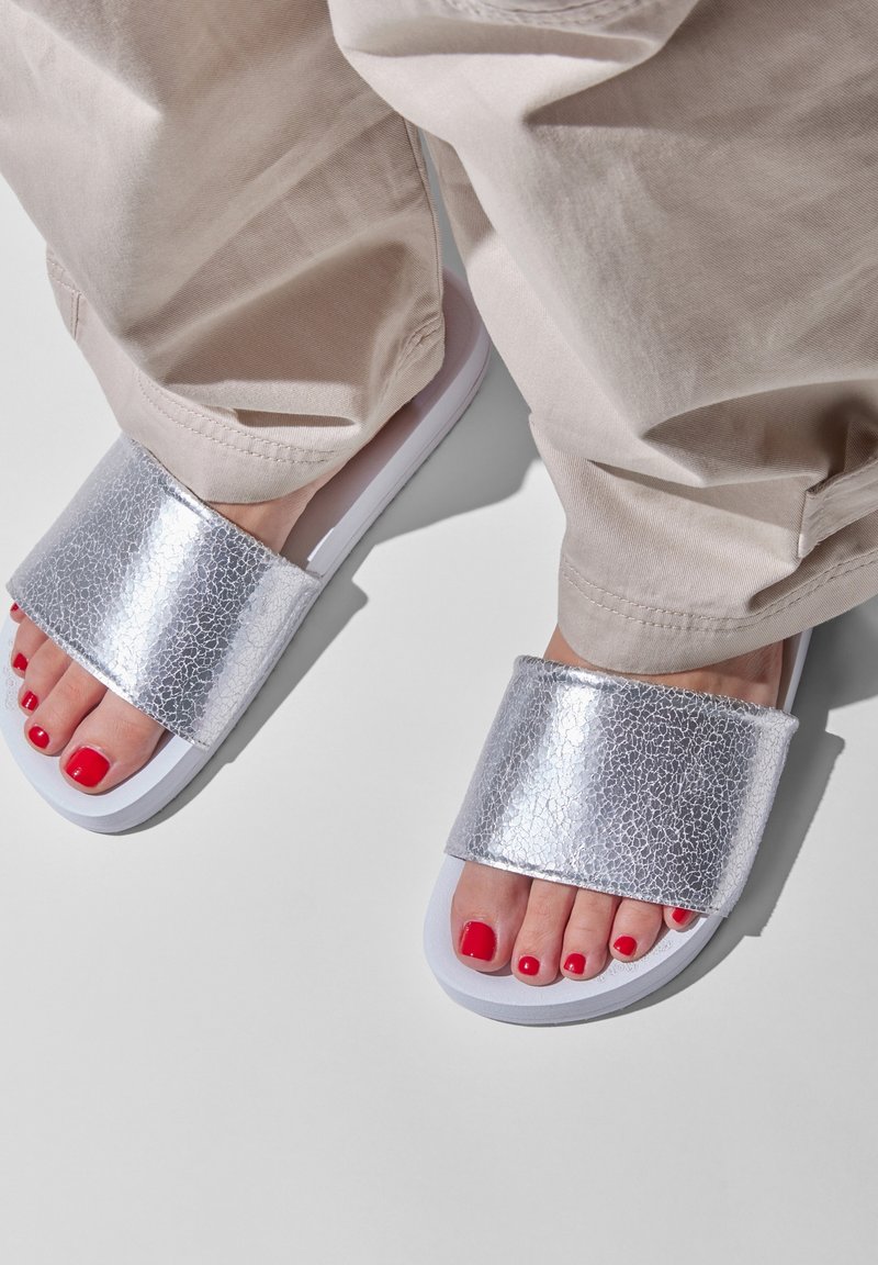 Silver textured slide sandals with a wide strap and a white sole, paired with light beige pants and red-painted toenails.