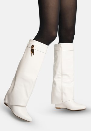 Hvide knæhøje kilehælstøvler med sidelåse og guld hængelåsdetaljer båret med sorte gennemsigtige strømpebukser på en lys baggrund.