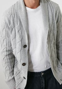 Grå kabelstickad cardigan med sjalkrage, två mörka knappar och ribbade ärmslut, bärs över en vit t-shirt och mörka jeans.