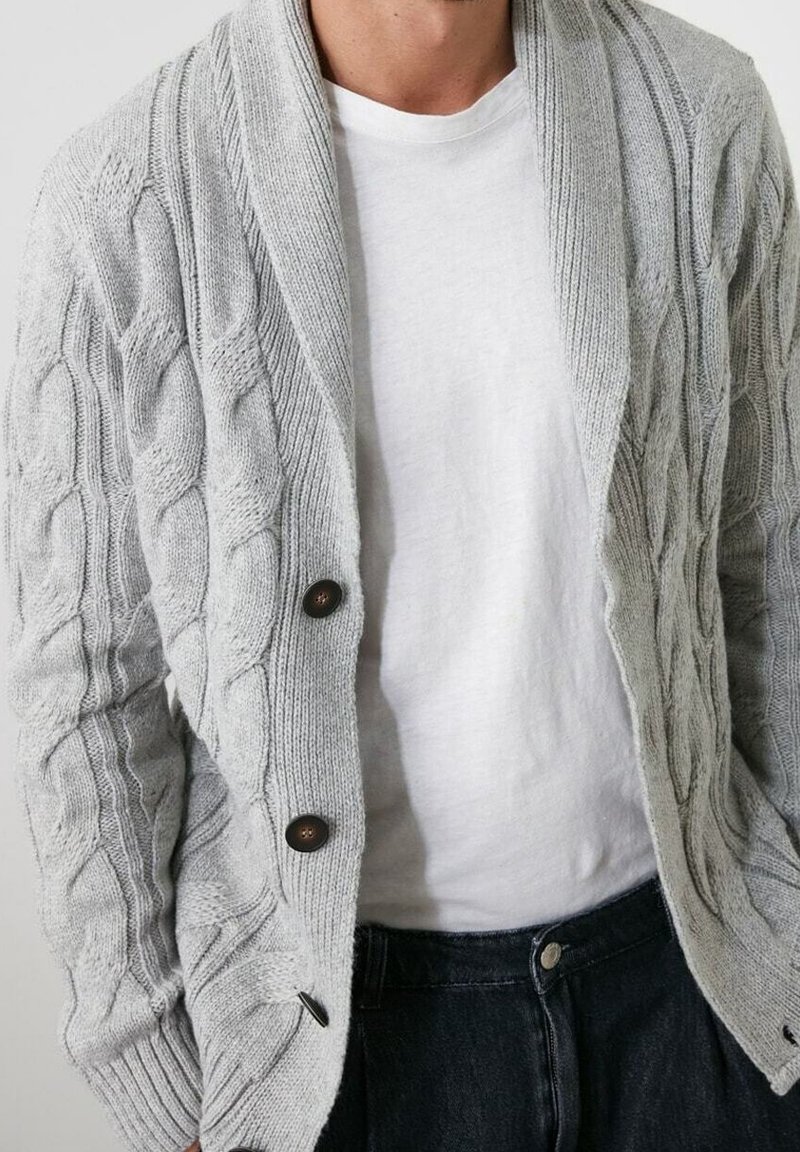 Grå kabelstickad cardigan med sjalkrage, två mörka knappar och ribbade ärmslut, bärs över en vit t-shirt och mörka jeans.