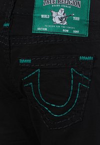 Bolsillo trasero de jeans negros con costuras en tonos verde azulado y una etiqueta verde que muestra un Buda tocando la guitarra, junto al texto "True Religion Brand Jeans".