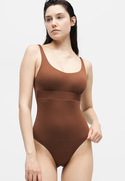 Γυναίκα με μακριά σκούρα μαλλιά, που φοράει καφέ ολόσωμο μαγιό, κοιτάζει ελαφρώς στο πλάι κρατώντας το τελείωμα του μαγιό με το ένα χέρι.