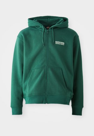 Corta-vento verde com fecho, feito de um tecido macio, com um bolso frontal, cordões e um logótipo no lado superior esquerdo.
