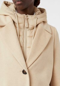 Cappotto in misto lana beige con revers a punta, chiusura con un bottone, sopra un puffy giubbotto trapuntato beige con zip visibile e cappuccio.