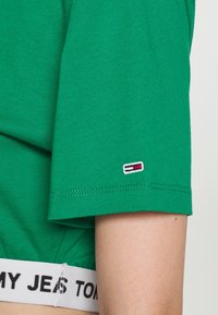 Zelené bavlněné tričko s krátkými rukávy, zdobené malým vyšitým logem v červené, bílé a modré barvě na levém rukávu a bílým páskem se značkou.