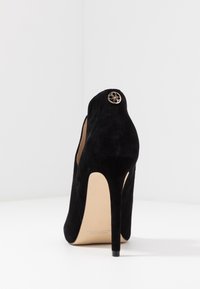 Musta mokkanahkainen korkokenkä, jossa on terävä varvas, koristeellinen kultakuvioinen yksityiskohta takana ja kapea stiletto-korko.