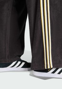 Black track pants with three yellow and white stripes on the sides, paired with black and white sneakers featuring a suede texture and white soles.