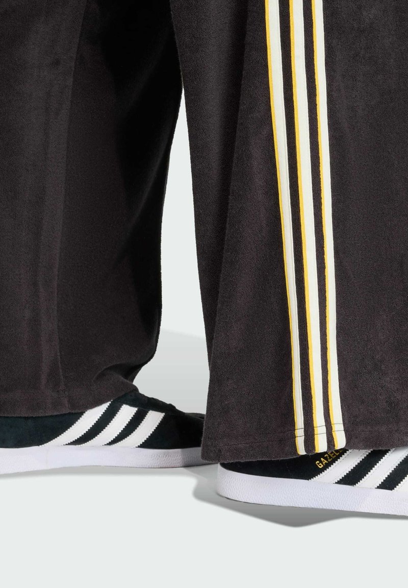 Pantaloni sportivi neri con tre strisce gialle e bianche sui lati, abbinati a sneakers nere e bianche con texture in suede e suole bianche.
