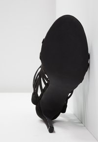 Chaussure noire à talon haut avec dessus à brides, vue de la semelle, placée contre un mur blanc sur une surface blanche.