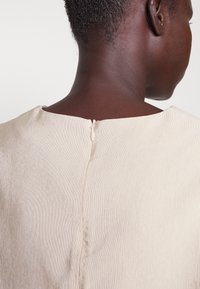 Beige-vaatekappale, jossa pystysuora ribbineulos, takana hillitty vetoketjusuljenta ja pyöreä pääntie.